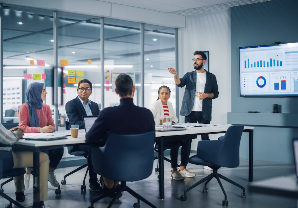 Diverse Modern Office: Businessman Leads Business Meeting with Managers, Talks, uses Presentation TV with Statistics, Infographics. Digital Entrepreneurs Work on e-Commerce Project.